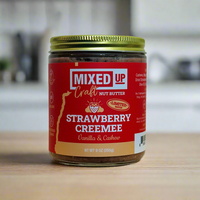 A glass jar of 'Mixed Up Craft Nut Butter' Strawberry Creemee flavor, with a red label and a yellow lid, placed on a kitchen countertop.